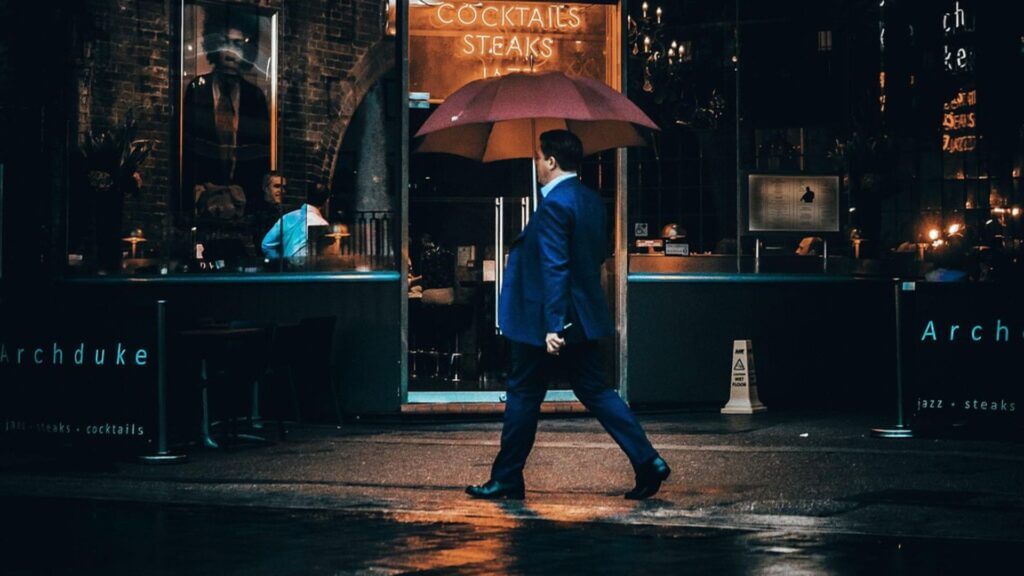 a man in blue suit walking with an pink umbrella on a wet street